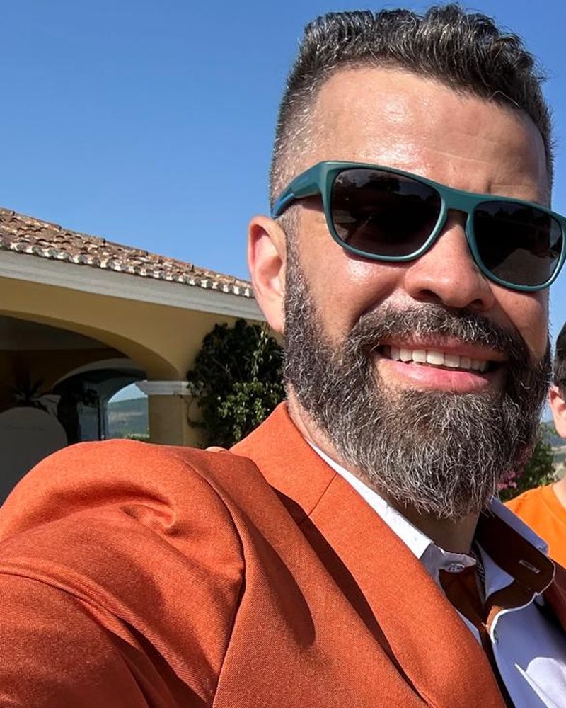 Guga Brasa de óculos escuros e blazer cor de laranja, a sorrir, num casamento em Portugal